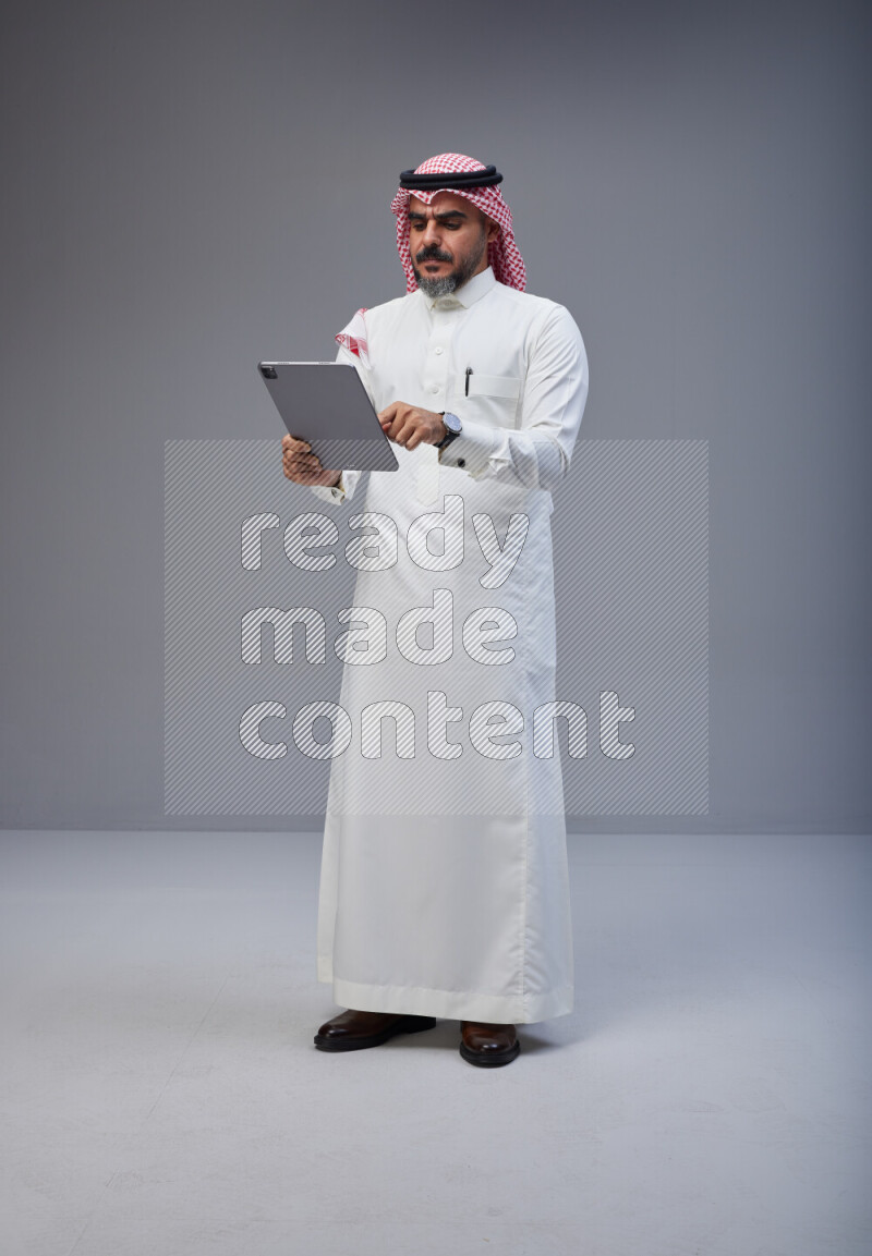 Saudi man Wearing Thob and red Shomag standing working on tablet on Gray background