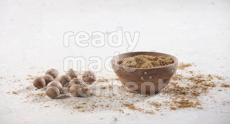 A wooden bowl full of nutmeg powder with whole seeds and sprinkled powder beside it on a textured white flooring