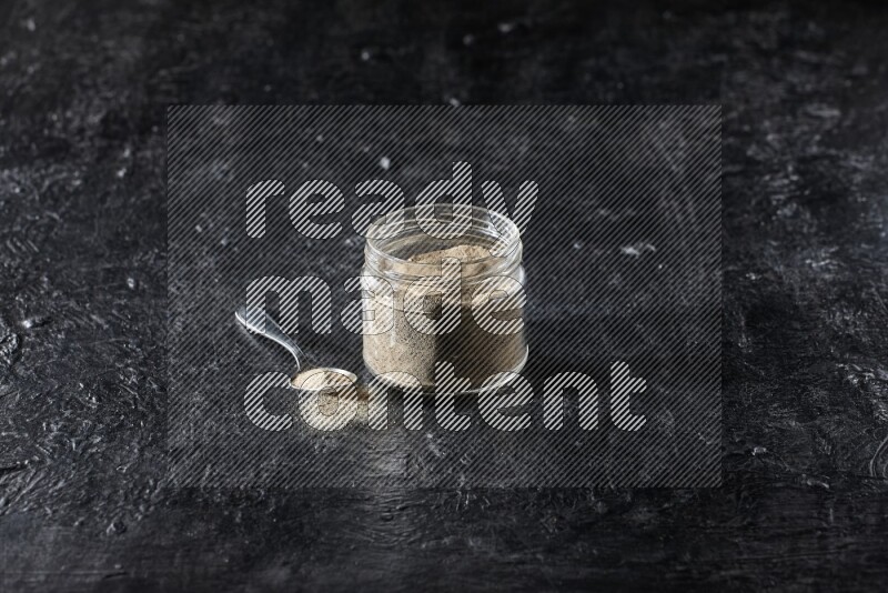 A glass jar and a metal spoon full of white pepper powder on textured black flooring