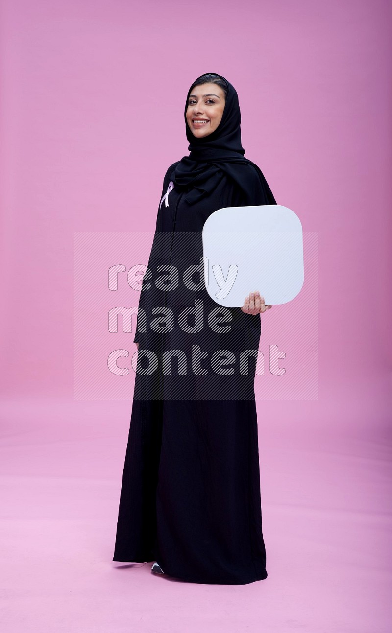 Saudi woman wearing pink ribbon on Abaya standing holding social media on pink background