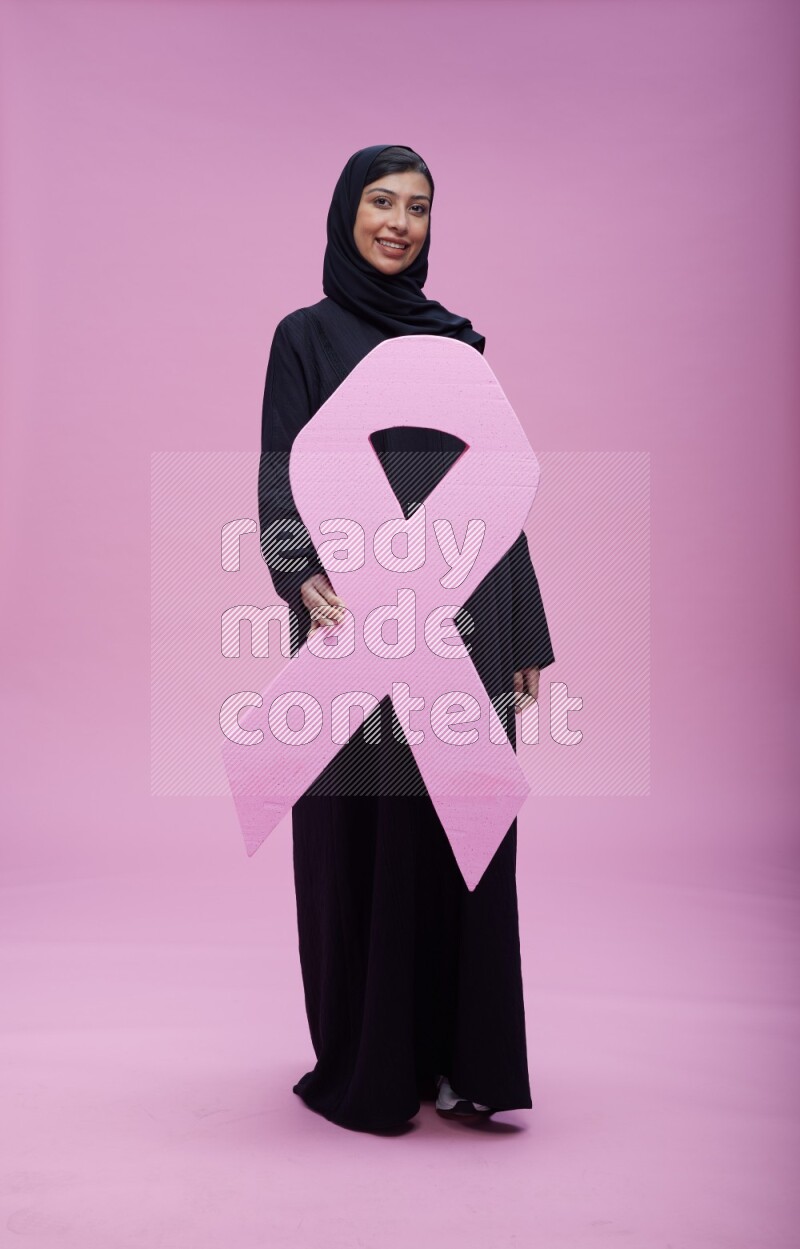 Saudi woman wearing pink ribbon on abaya standing holding awareness ribbon on plain pink background