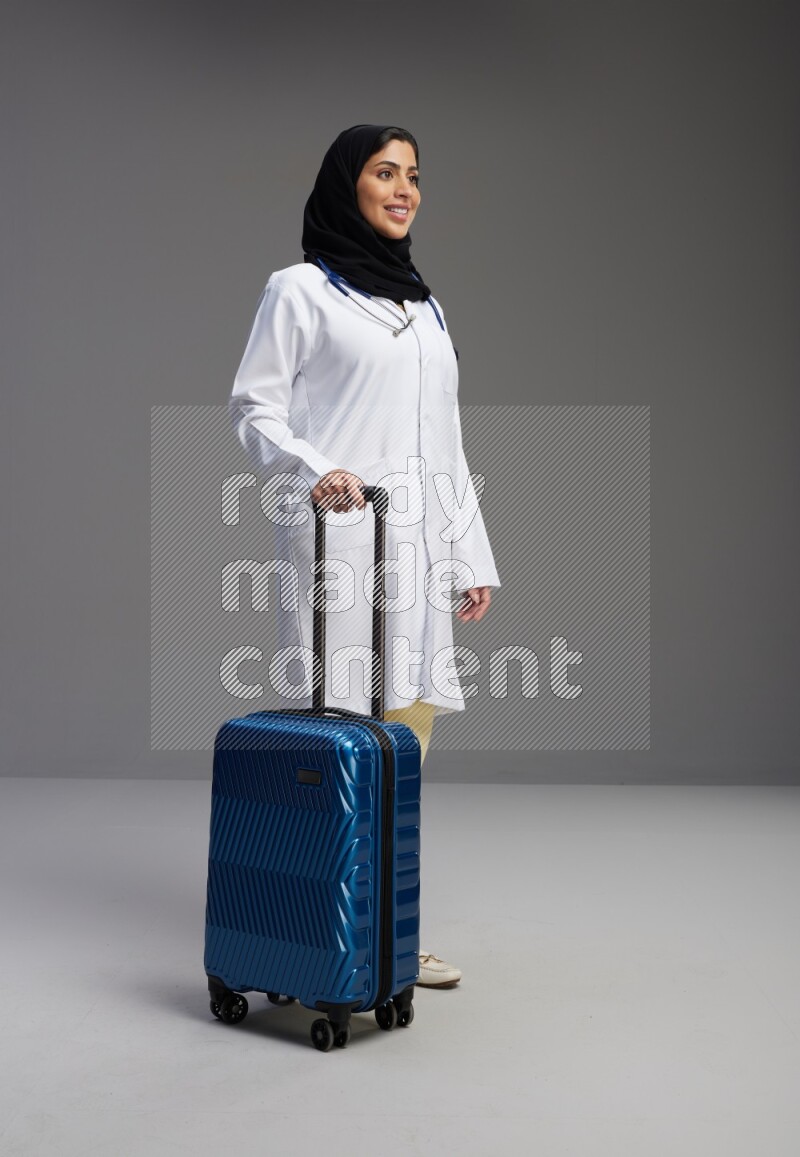 Saudi woman wearing lab coat with stethoscope standing holding Travel bag on Gray background