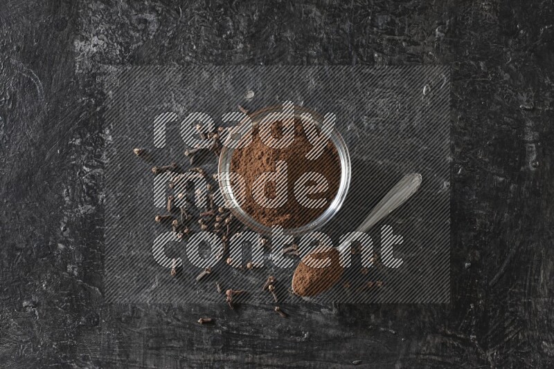 وعاء زجاجي وملعقة معدنية ممتلئان ببودرة القرنفل مع حبوب القرنفل بجانبهما على خلفية سوداء