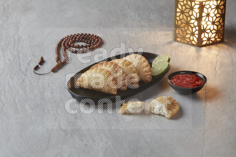 Four fried sambosas in an oval shaped black plate, beside a cut cheese sambosa, a brown misbaha and a golden lantern on a gray background