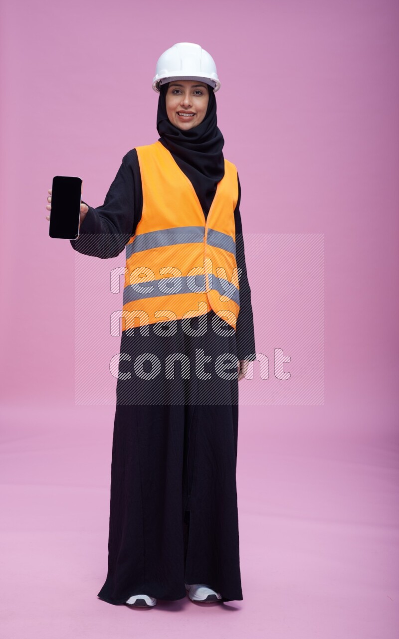 Saudi woman wearing Abaya with engineer vest and helmet standing showing phone to camera on pink background