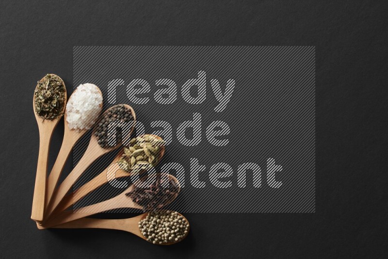 wooden spoons filled with white peppers, cloves, cardamom, salt, black peppers and basil on black flooring