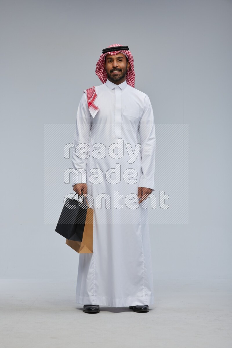Saudi man Wearing Thob and shomag standing holding shopping bag on Gray background