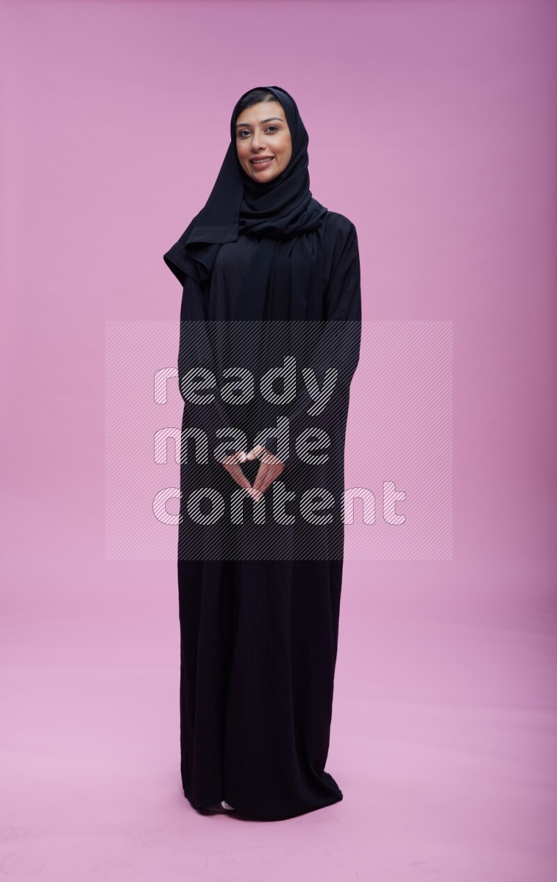 Saudi woman wearing Abaya standing interacting with the camera on pink background