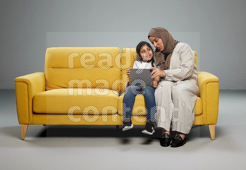 A girl with her mother on yellow sofa and watching on iPad on gray background