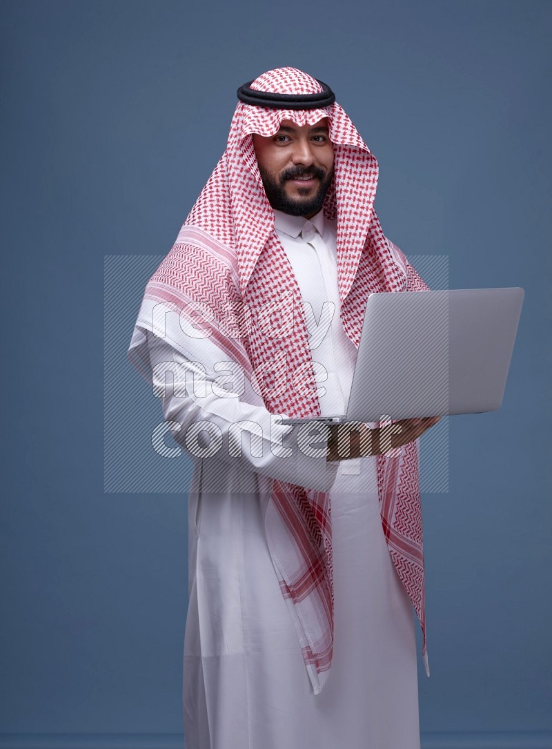A man standing  with a laptop on Blue Background wearing Saudi Thob and Shomag