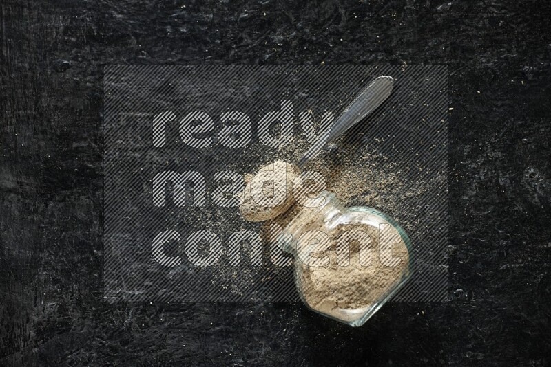 A flipped glass spice jar and metal spoon full of cardamom powder and powder spilled out of it on textured black flooring