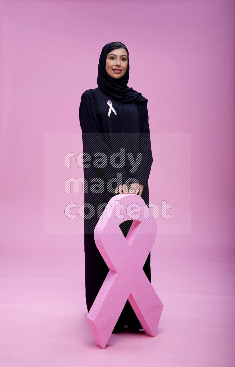 Saudi woman wearing pink ribbon on abaya standing holding awareness ribbon on plain pink background