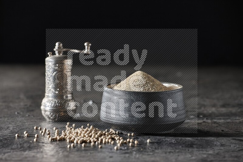 Black pottery bowl full of white pepper powder with pepper beads and metal pepper grinder on textured black flooring