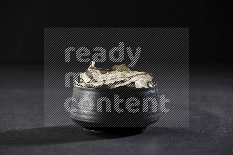 A black pottery bowl full of dried bay leaves on black flooring