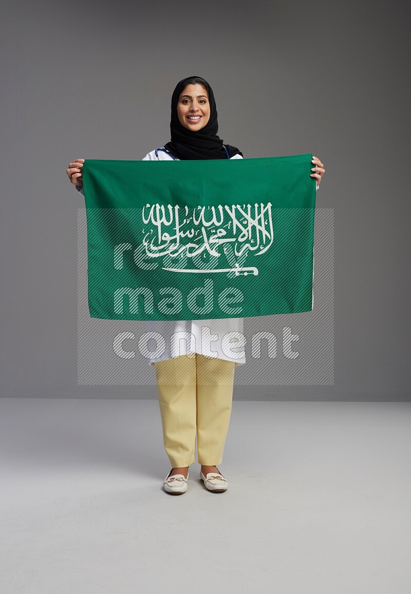 Saudi woman wearing lab coat with stethoscope standing holding Saudi flag on Gray background