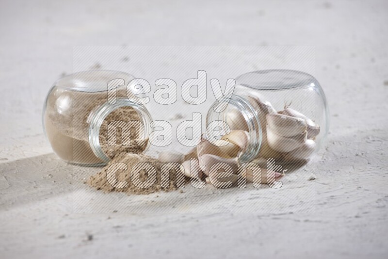 2 glass spice jars, one full of garlic powder and the other full of garlic cloves both flipped on a textured white flooring