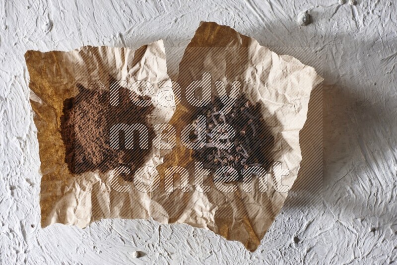 Cloves powder and cloves on 2 crumpled pieces of paper on a textured white flooring