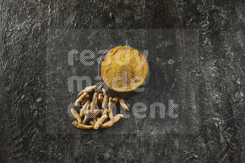 A black pottery bowl full of turmeric powder and dried turmeric whole fingers next of it on textured black flooring