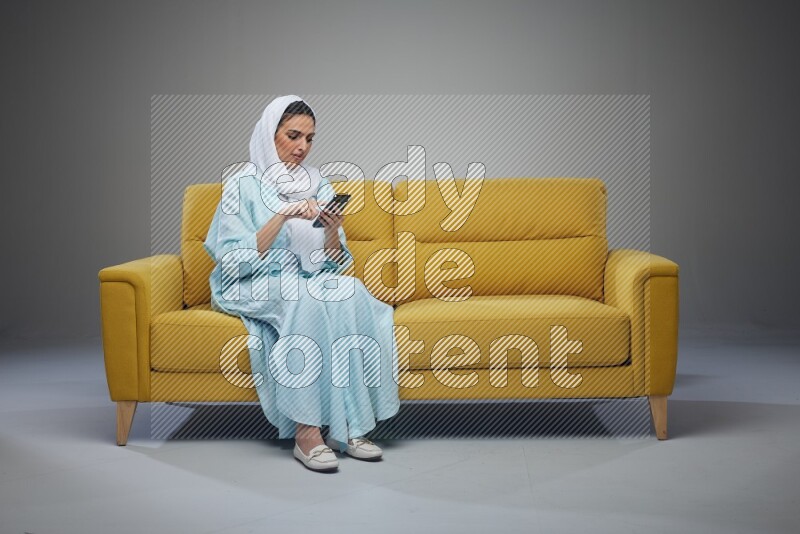 A Saudi woman wearing a light blue Abaya and white head scarf sitting on a yellow sofa and using her phone eye level on a grey background