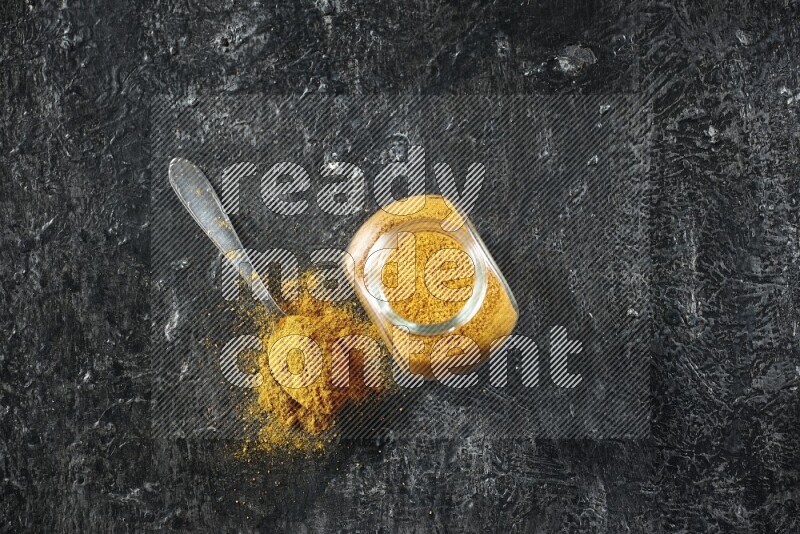 وعاء زجاجي للتوابل وملعقة معدنية ممتلئان ببودرة الكركم على خلفية سوداء