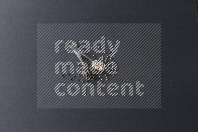 ملعقتان معدنيتان ممتلئتان بحبوب الفلفل الأسود والأبيض مع حبوب متناثرة على أرضية سوداء