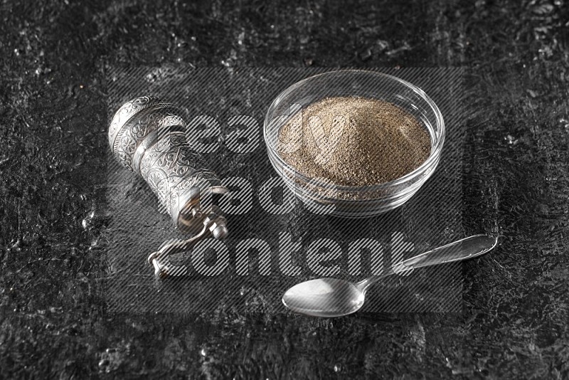 A glass bowl full of black pepper powder with a turkish metal pepper grinder and a metal spoon on textured black flooring