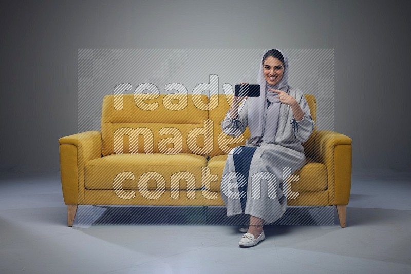 A Saudi woman wearing a light gray Abaya and head scarf setting on a yellow sofa and holding her phone while showing it's screen eye level on a grey background