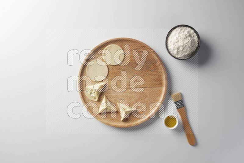 two closed sambosas and one open sambosa filled with cheese while flour, and oil with oil brush aside in a wooden dish on a white background
