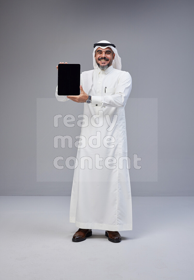 Saudi man Wearing Thob and white Shomag standing showing tablet to camera on Gray background