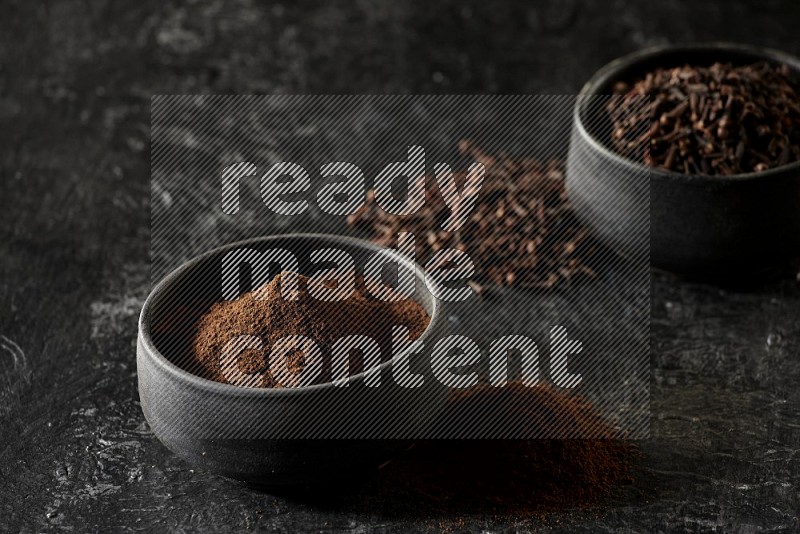 2 Black pottery bowls full of cloves and the other full of cloves powder on textured black flooring