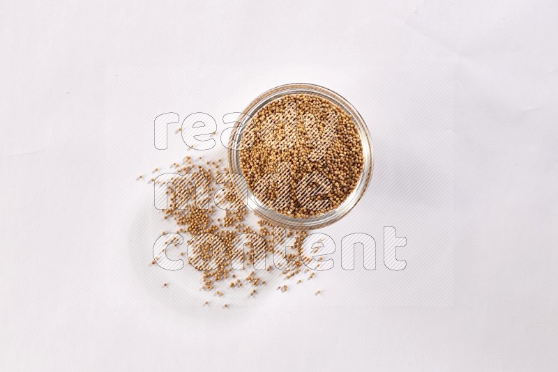 A glass bowl full of mustard seeds and more seeds spread on a white flooring in different angles