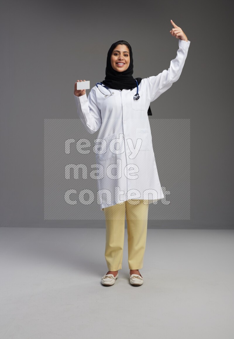 Saudi woman wearing lab coat with stethoscope standing holding ATM card on Gray background
