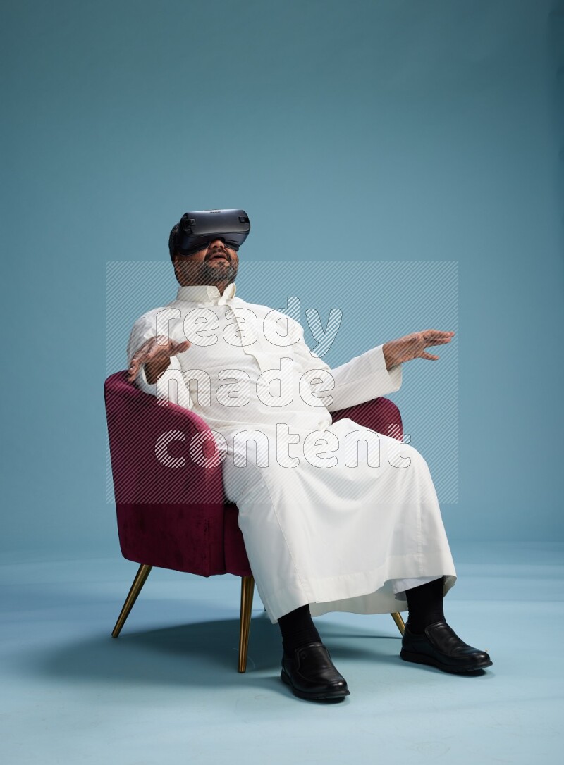 Saudi Man without shimag sitting on chair  gaming with VR on blue background