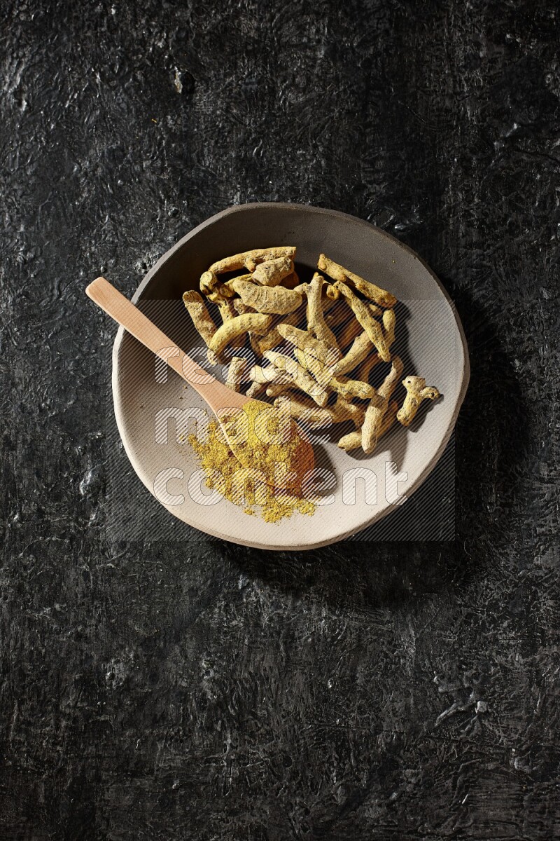 A plate filled with dried turmeric fingers and a wooden spoon full of turmeric powder on a textured black flooring