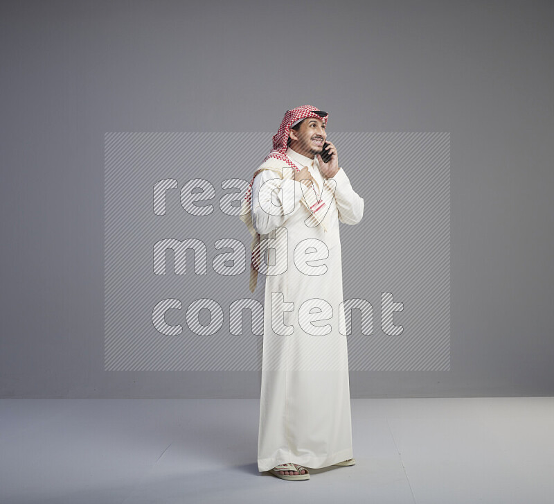 A Saudi man standing wearing thob and red shomag talking on phone on gray background