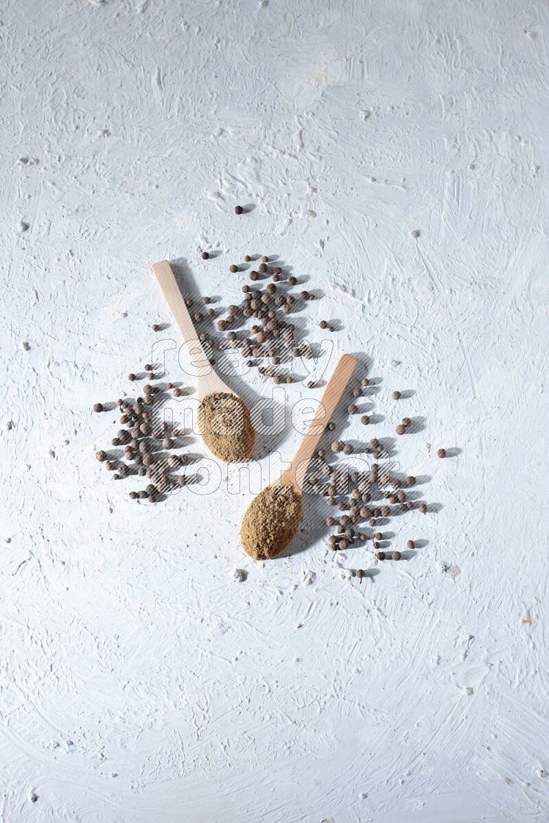 Wooden spoons full of allspice powder and allspice whole balls spreaded on a textured white flooring