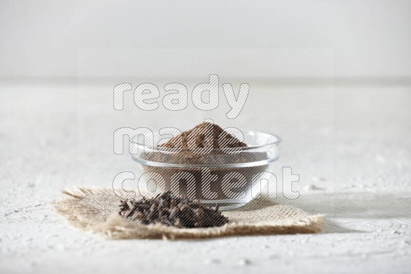 A glass bowl full of cloves powder and cloves grains on a burlap piece on a white flooring