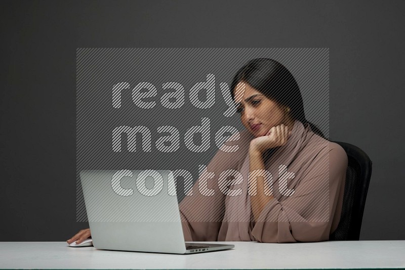 A Saudi woman Sitting on her desk on a Gray Background wearing Brown Abaya