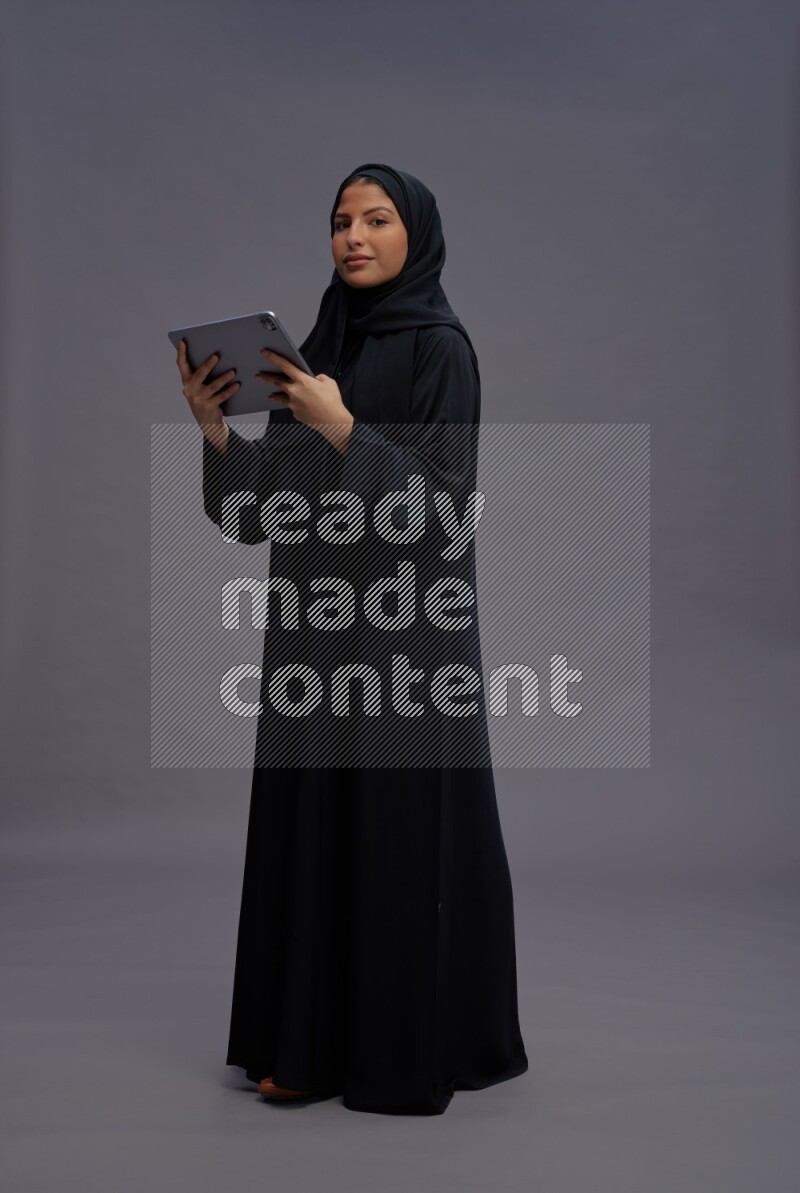 Saudi woman wearing Abaya standing working on tablet on gray background