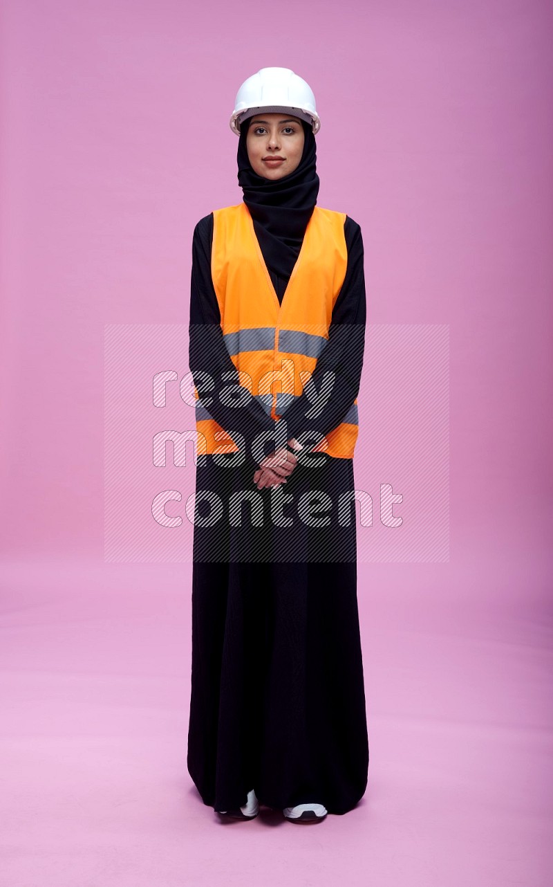 Saudi woman wearing Abaya with engineer vest and helmet standing interacting with the camera on pink background