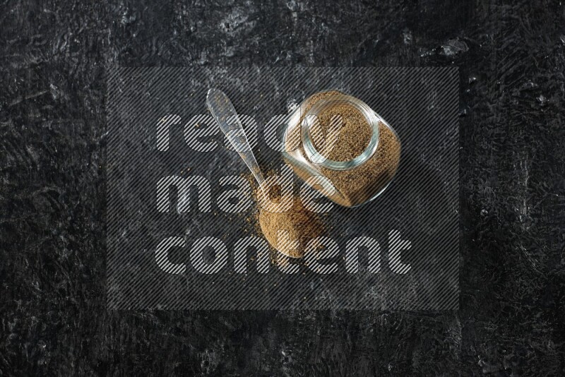 A glass spice jar and a metal spoon full of cumin powder on a textured black flooring