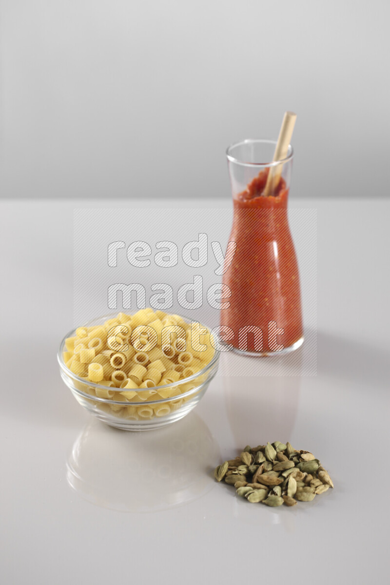 Raw pasta with tomatoe pasta with different ingredients such as cherry tomatoes, basil, garlic, bay laurel, cardamom, white pepper, black pepper, red chilis and wheat stalks on light grey background