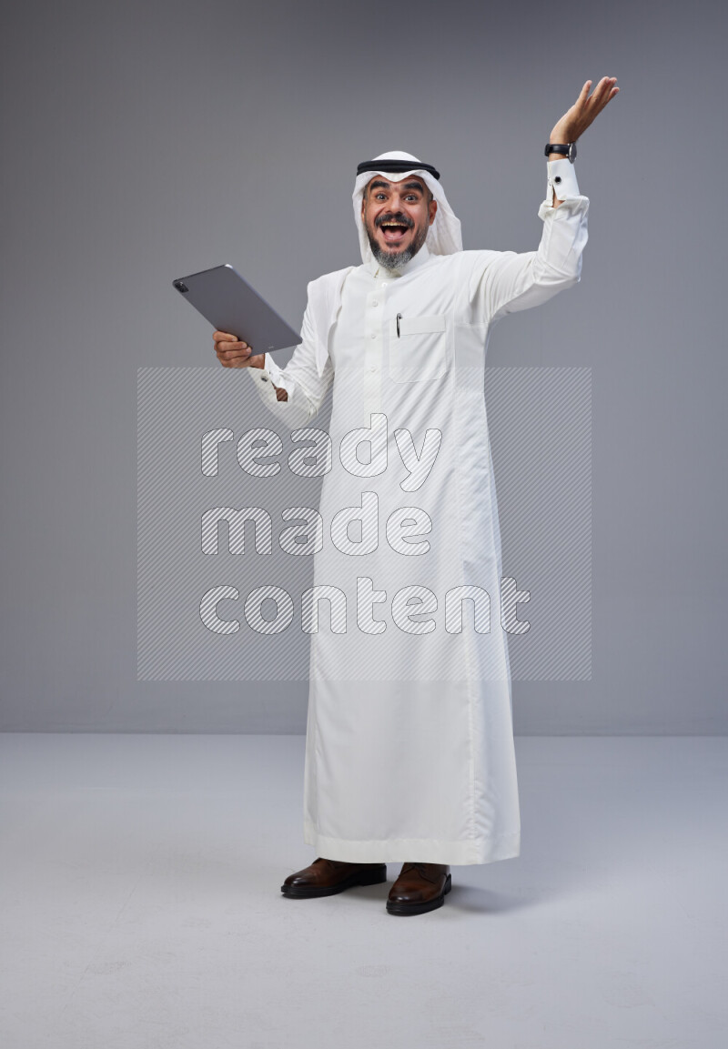 Saudi man Wearing Thob and white Shomag standing working on tablet on Gray background