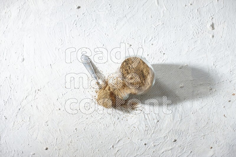 وعاء زجاجي للتوابل مقلوب وملعقة معدنية كلاهما ممتلئ ببودرة الكمون والبودرة تناثرت منه على خلفية بيضاء