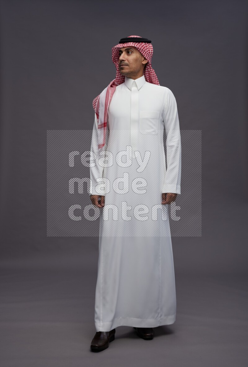 Saudi man wearing thob and shomag standing interacting with the camera on gray background