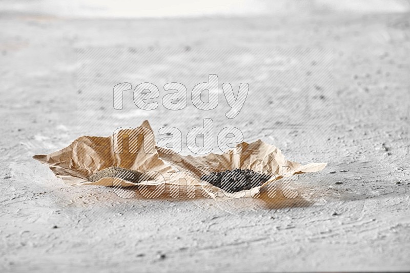 Black pepper and black pepper powder on 2 crumpled pieces of craft paper on a textured white flooring