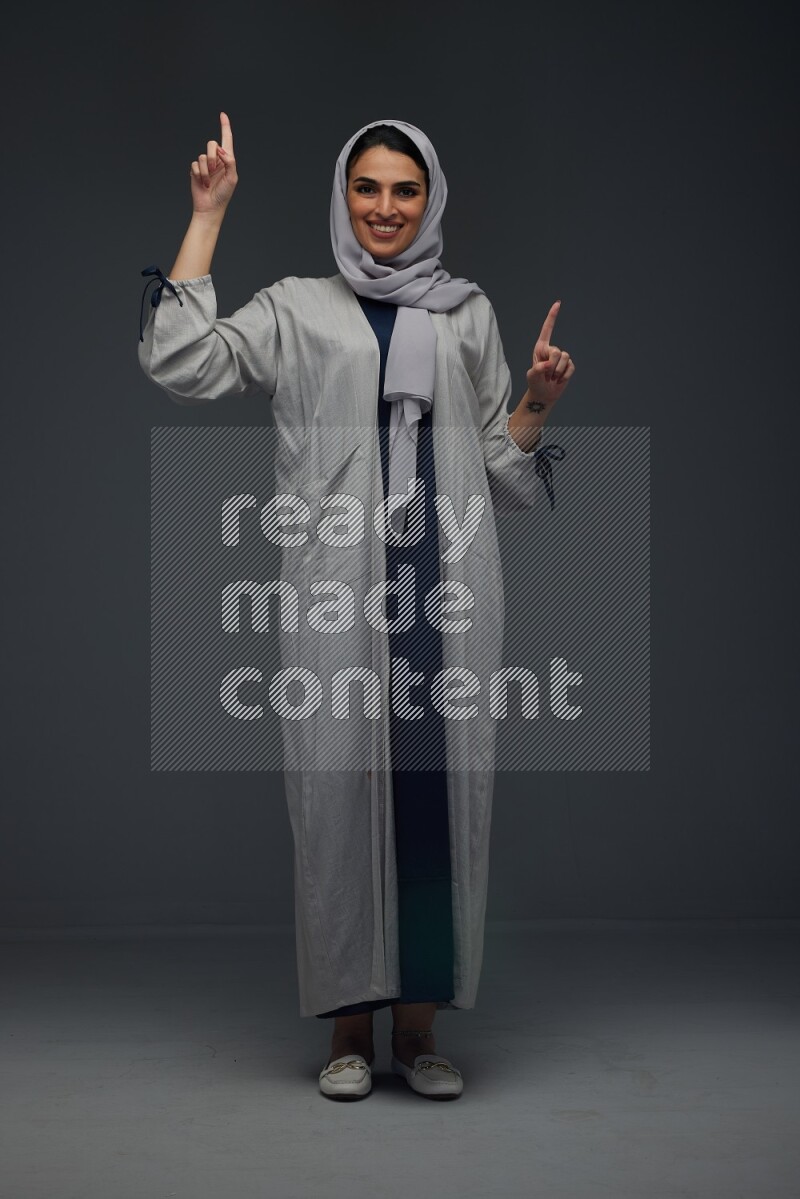 A Saudi woman wearing a light gray Abaya and head scarf standing and making multi poses eye level on a grey background