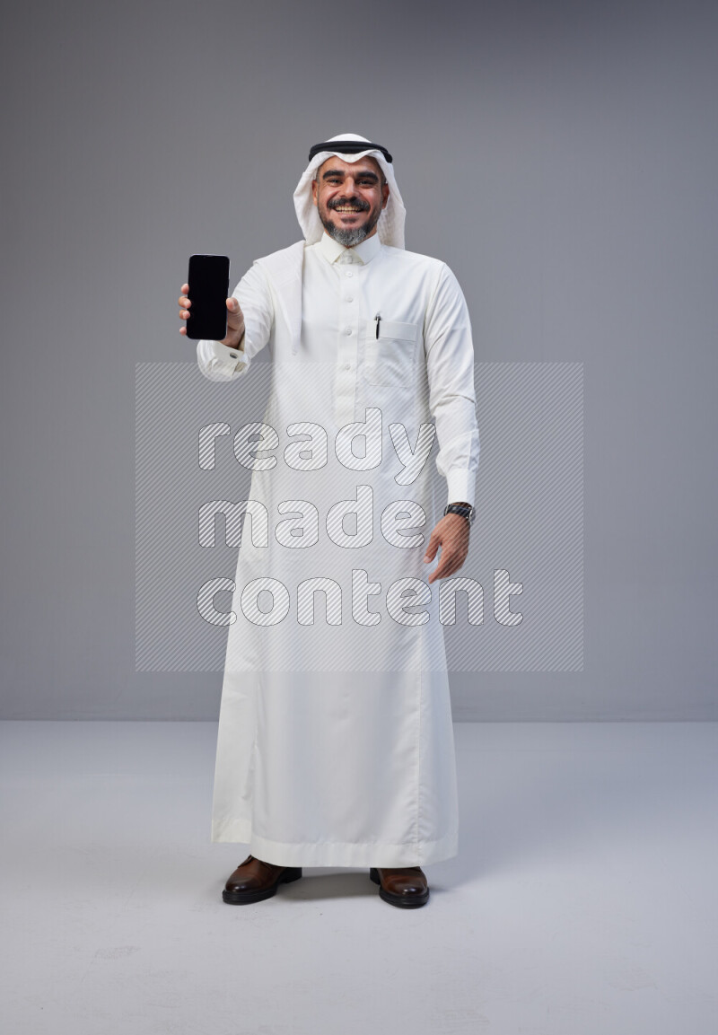 Saudi man Wearing Thob and white Shomag standing showing phone to camera on Gray background