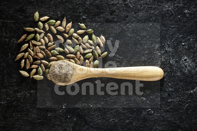 A wooden spoon full of cardamom powder and cardamom seeds next of it on textured black flooring