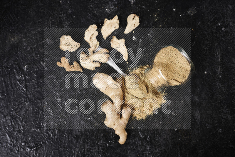 A glass jar full of ground ginger powder flipped with some spilling powder on black background
